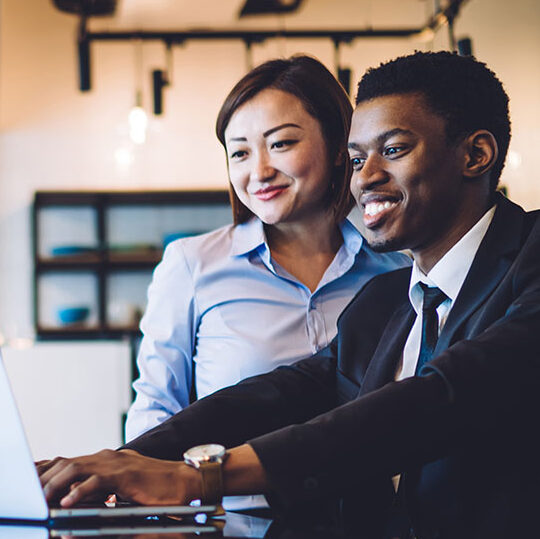 African American businessman with toothy smile in black suit sitting at table with Asian cheerful female colleague and working with computer